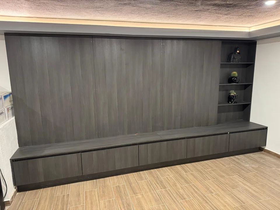 Centro de TV de acabado gris con panel posterior, mueble bajo, nichos laterales e iluminación superior cálida.