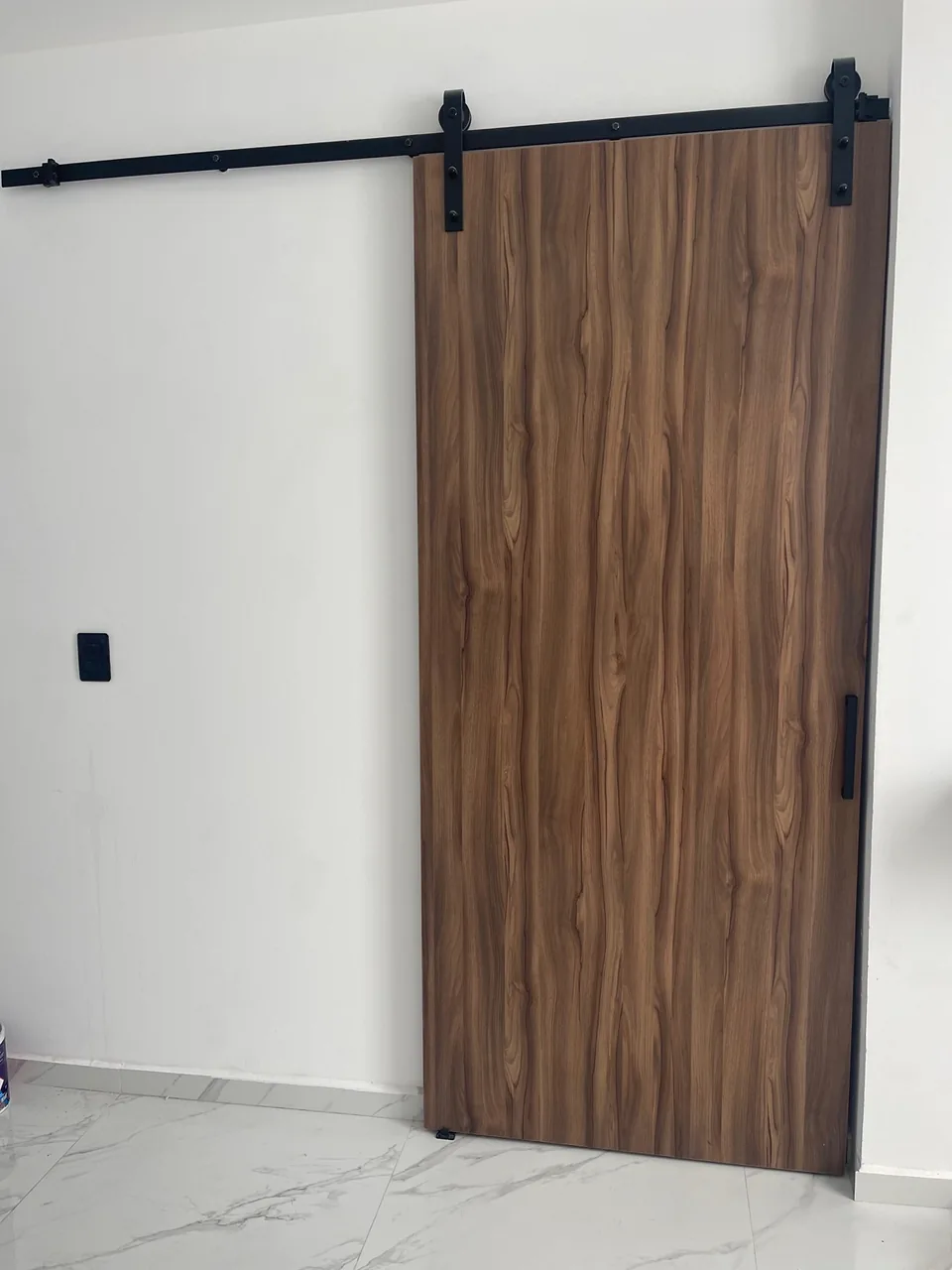 Puerta corrediza de veta vertical en tono madera con riel y herrajes negros visibles.