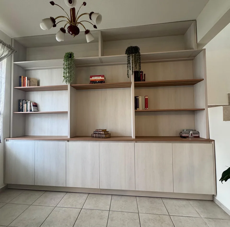 Librero de piso a techo en tonos claros, con repisas abiertas y puertas inferiores, libros y plantas.