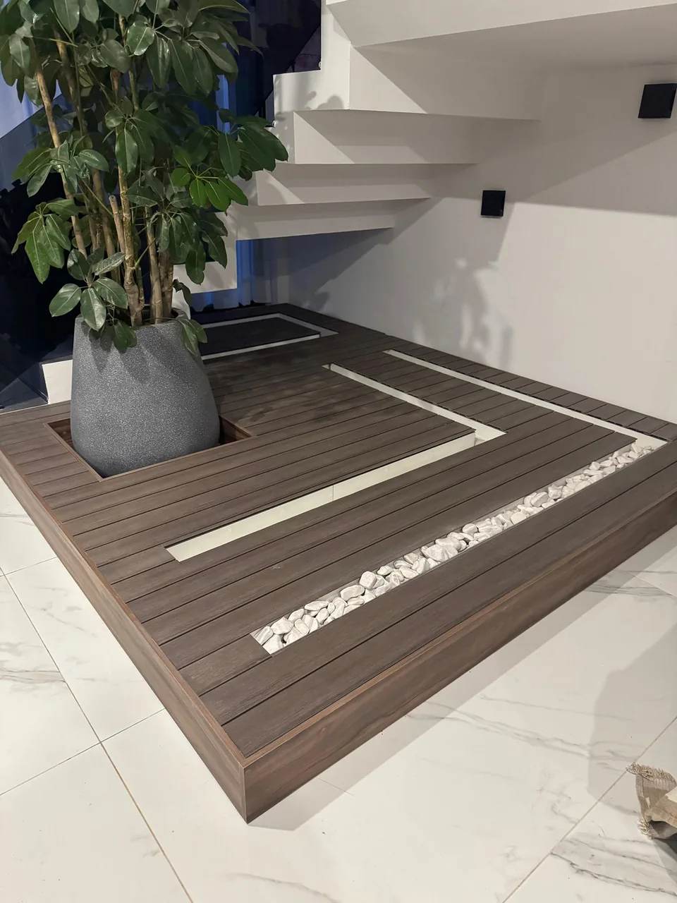 Plataforma de acabado en tono madera bajo una escalera, con maceta, franjas claras y piedras decorativas.