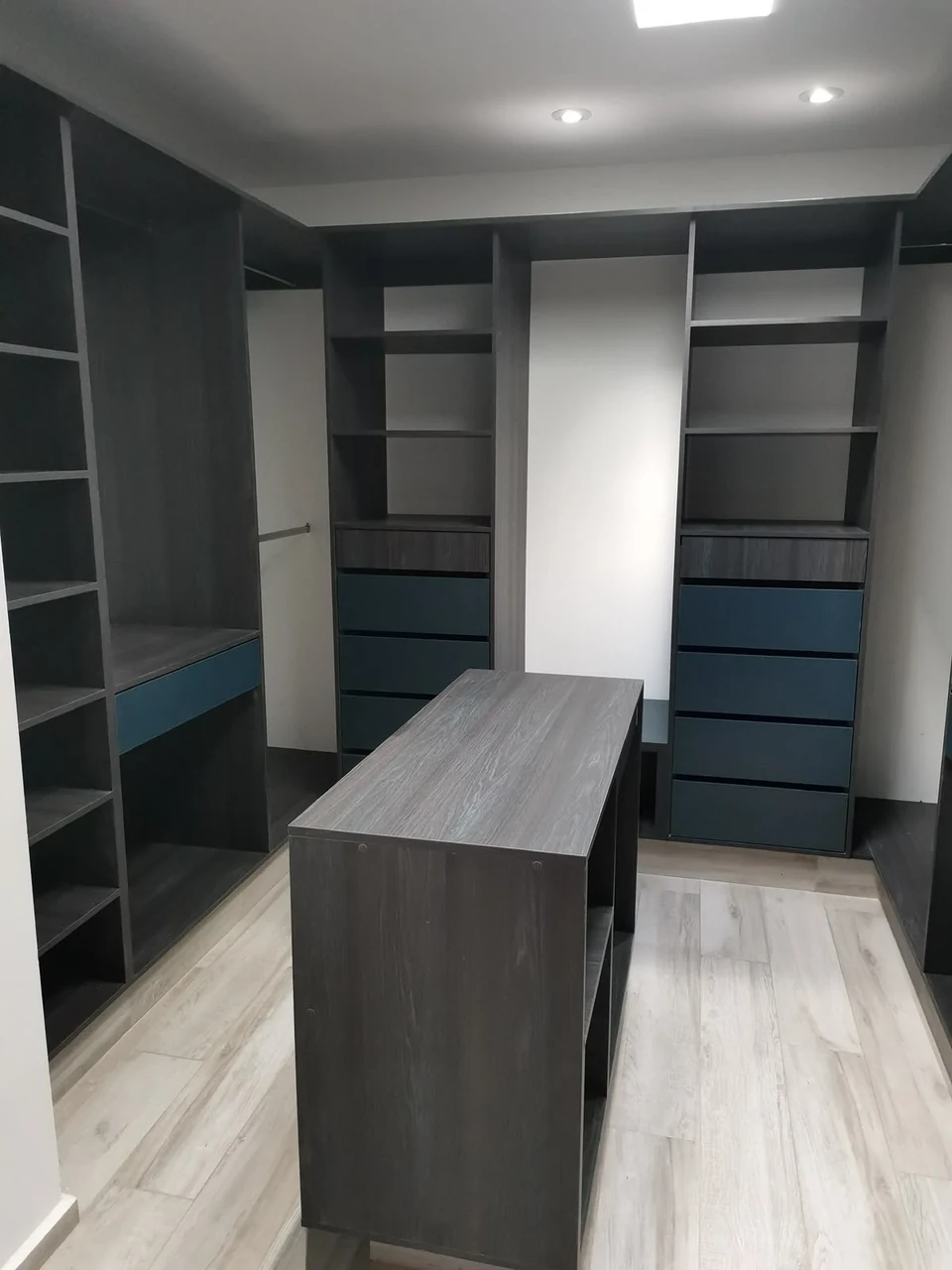 Vestidor con módulos en tono madera oscuro, cajones azules e isla central.